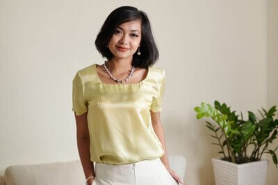 Portrait of positive young Asian woman in satin blouse standing in bright room
